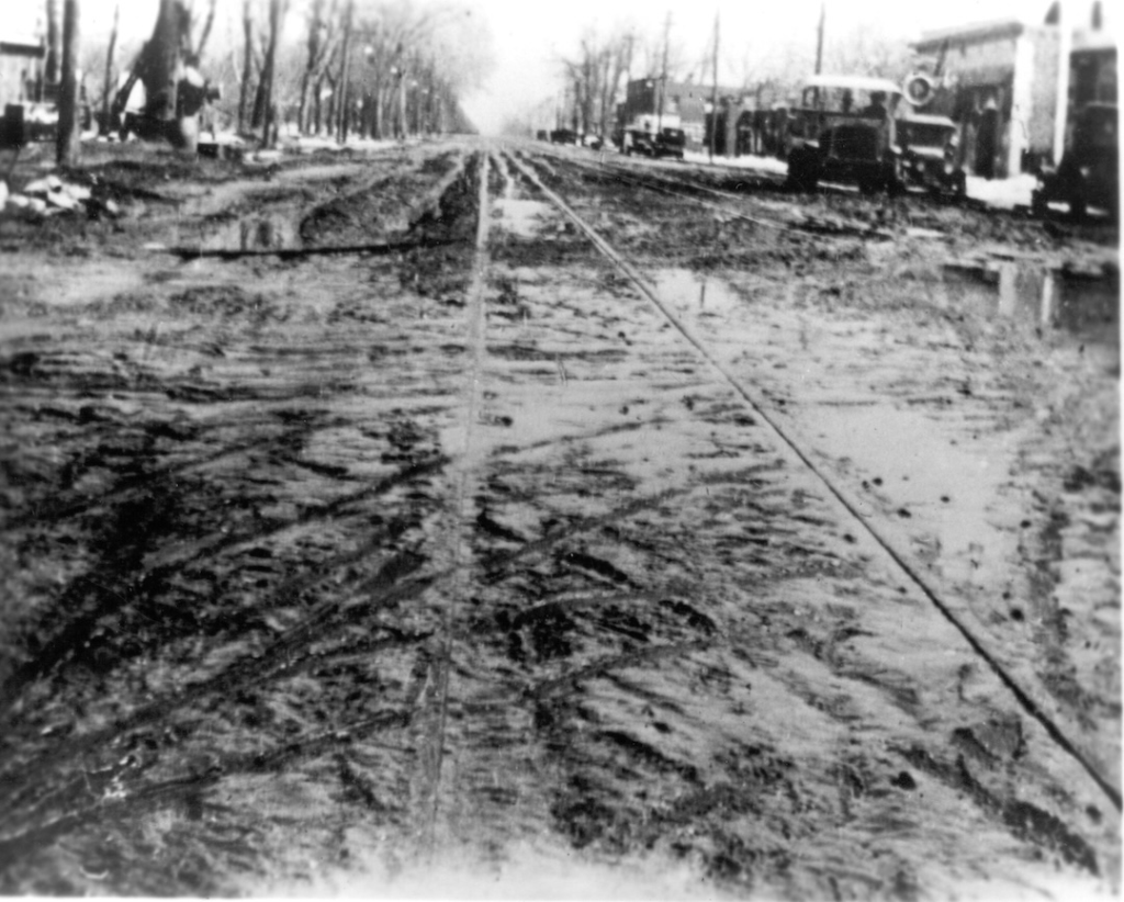 A black-and-white photo of a muddy, rutted street with visible streetcar tracks; vehicles and leafless trees line both sides, evoking the historic ambiance of BookCliff Vineyards, a pioneer in Colorado wine.